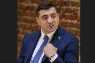 Un bărbat cu părul scurt, purtând sacou albastru, cămașă albă și cravată albastră, stă în fața unui perete de cărămidă roșie.