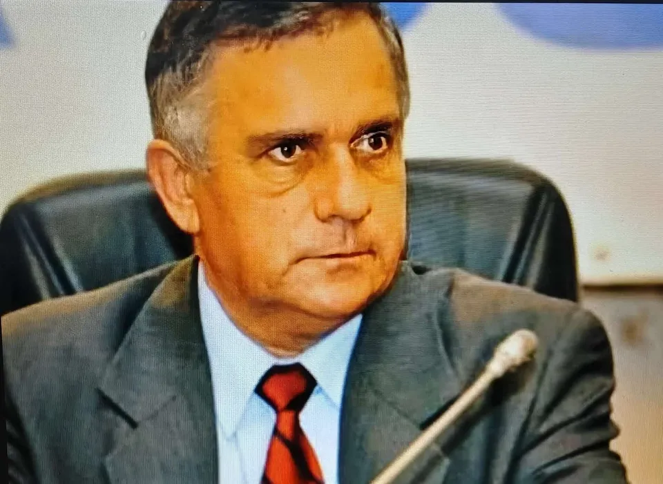 Un bărbat cu părul scurt și cămășă albastră, purtând o vestă gri și o cravată roșie, stă așezat într-un scaun de birou negru.