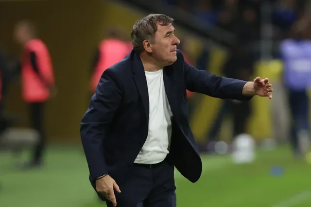 Un bărbat într-un sacou închis la culoare și tricou alb stă pe un teren de fotbal cu brațul întins spre lateral.