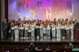 Un grup mare de copii și adulți stau pe o scenă iluminată, mulți dintre ei ținând diplome albe în mâini.