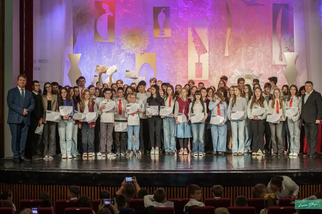 Un grup mare de copii și adulți stau pe o scenă iluminată, mulți dintre ei ținând diplome albe în mâini.