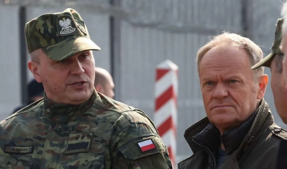 Un bărbat în uniformă militară de camuflaj și un bărbat într-o jachetă închisă la culoare stau unul lângă celălalt în exterior.
