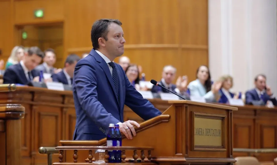 Un bărbat într-un costum albastru stă în spatele unui podium de lemn într-o sală cu mese și oameni în fundal.