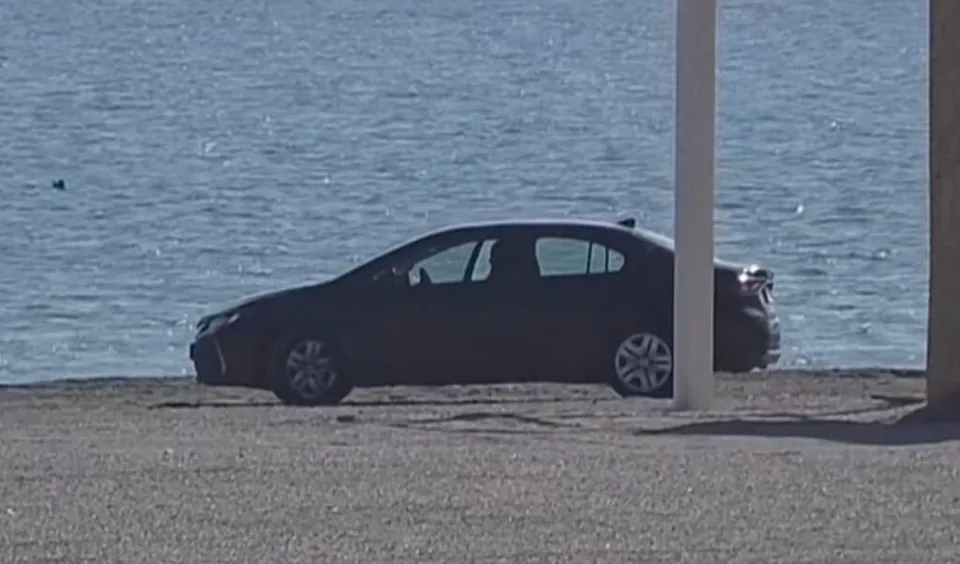 Un autoturism de culoare închisă este parcat pe o suprafață nisipoasă lângă o întindere de apă sub un cer luminos.