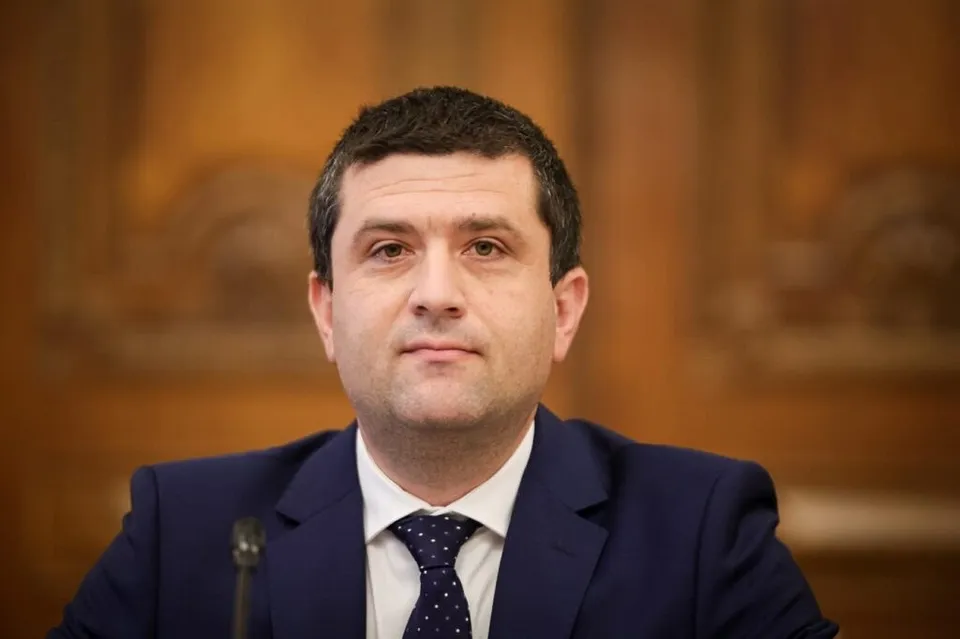 Un bărbat cu părul scurt și întunecat poartă un costum albastru, cămașă albă și cravată închisă la culoare în fața unui fundal lemnos.