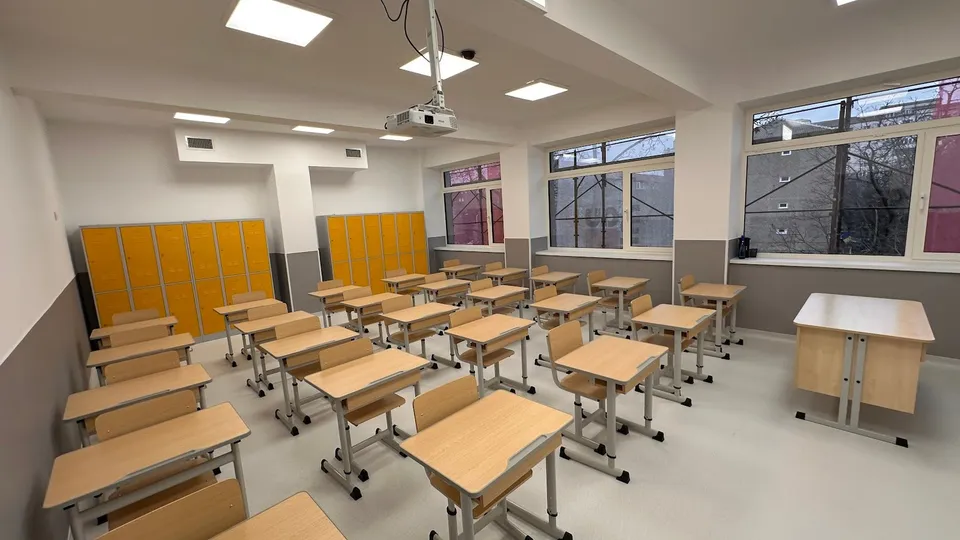 O sală de clasă cu mai multe birouri de lemn cu picioare metalice gri și ferestre mari pe un perete lateral.