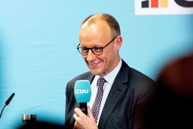 Un bărbat cu ochelari și cravată cu model punctat zâmbește în timp ce ține un microfon cu inscripția CDU.