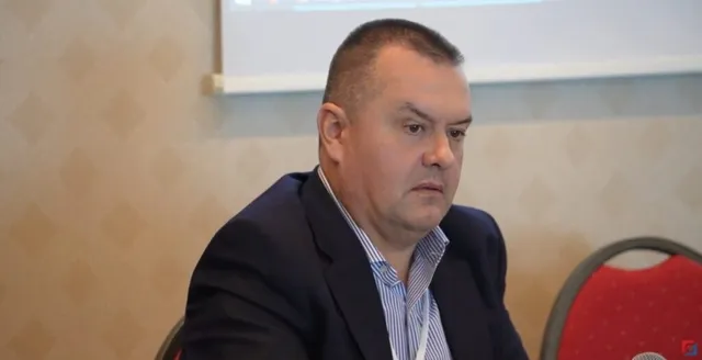 Un bărbat cu părul scurt, purtând sacou închis la culoare și cămașă cu dungi, stă așezat într-o încăpere cu un fundal de perete texturat.