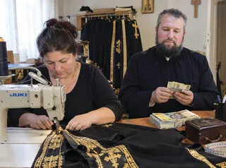 O femeie folosește o mașină de cusut pentru a lucra la un material negru cu broderii aurii, în timp ce un bărbat cu barbă privește.