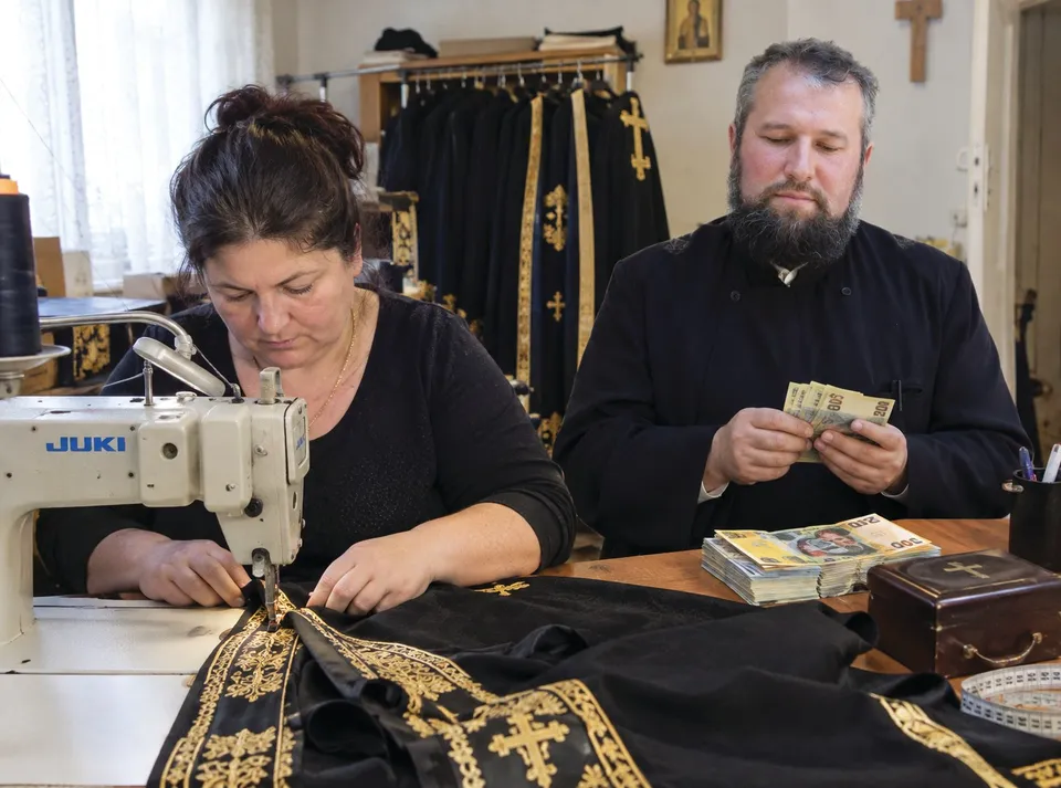 O femeie folosește o mașină de cusut pentru a lucra la un material negru cu broderii aurii, în timp ce un bărbat cu barbă privește.
