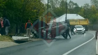 Un autoturism roșu este înclinat pe partea de lângă marginea drumului, lângă un cap de pod de beton, în prezența unor persoane.