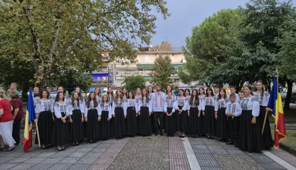 Un grup de copii și tineri îmbrăcați în costume tradiționale albe și negre stau în rând pe o esplanadă lângă două steaguri.