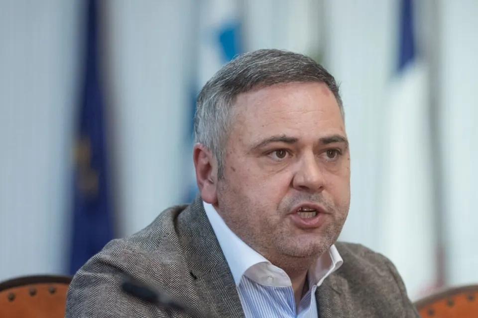 Un bărbat cu părul scurt și cămășă albastră sub un sacou maro stă cu gura ușor deschisă.