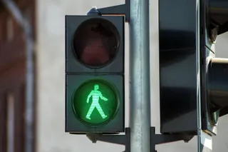 Un semafor de pietoni afișează o siluetă verde luminoasă reprezentând un om care merge pe un fundal circular verde în interiorul unei carcase negre.