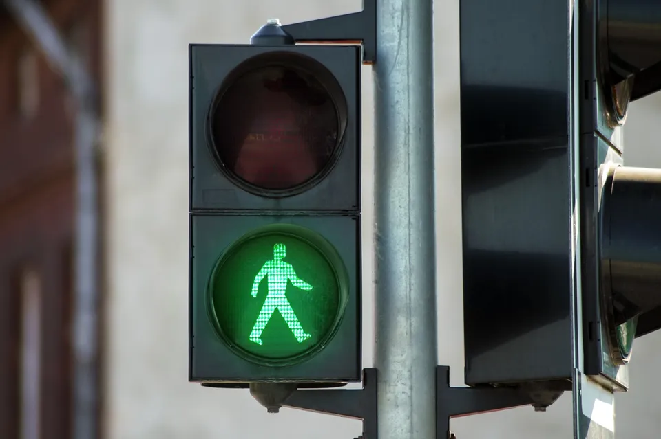 Un semafor de pietoni afișează o siluetă verde luminoasă reprezentând un om care merge pe un fundal circular verde în interiorul unei carcase negre.