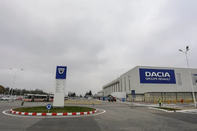 Imaginea prezintă o clădire modernă cu sigla "Dacia Groupe Renault" pe fațadă, un sens giratoriu și un panou informativ cu logo-ul Dacia în prim-plan.