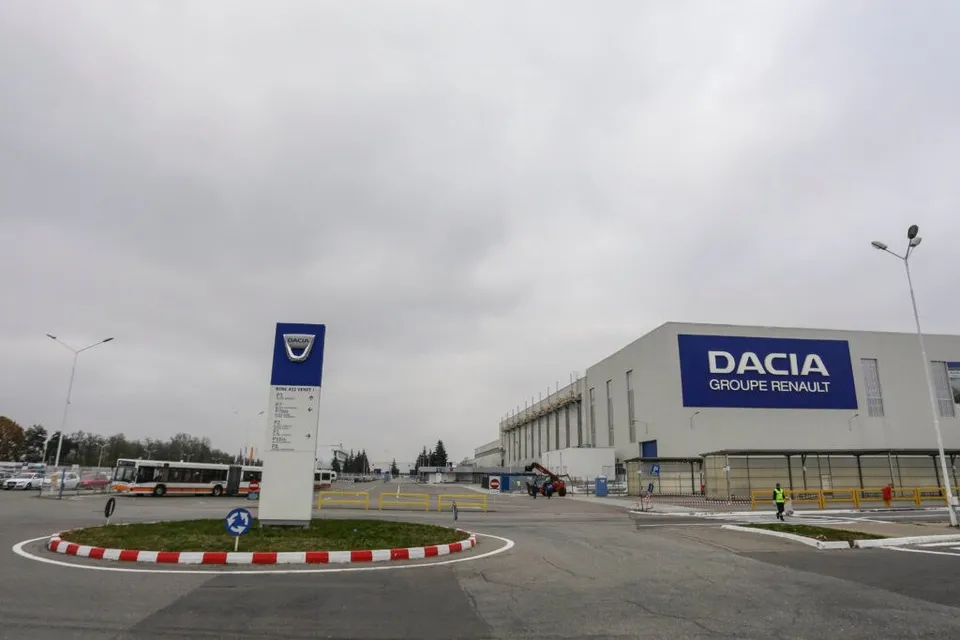 Imaginea prezintă o clădire modernă cu sigla "Dacia Groupe Renault" pe fațadă, un sens giratoriu și un panou informativ cu logo-ul Dacia în prim-plan.