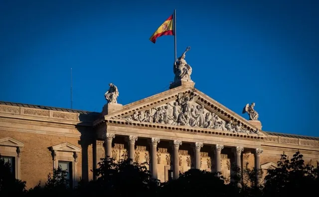 Un steag spaniol flutură pe un stârf deasupra unei clădiri cu coloane și sculpturi pe fațadă sub un cer albastru.