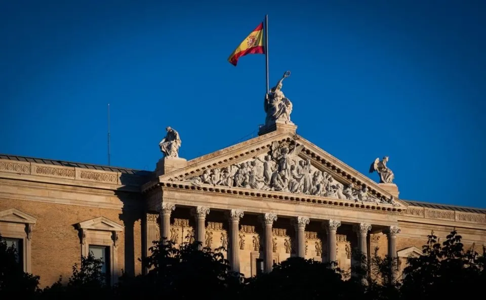 Un steag spaniol flutură pe un stârf deasupra unei clădiri cu coloane și sculpturi pe fațadă sub un cer albastru.