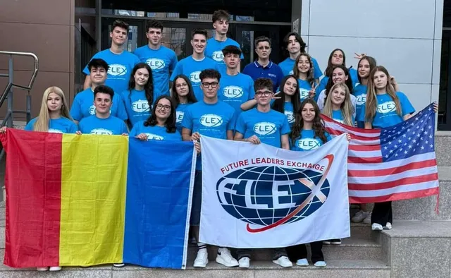 Un grup de tineri stă în rând, în fața unei clădiri, ținând un steag american și un steag alb cu un logo circular colorat.