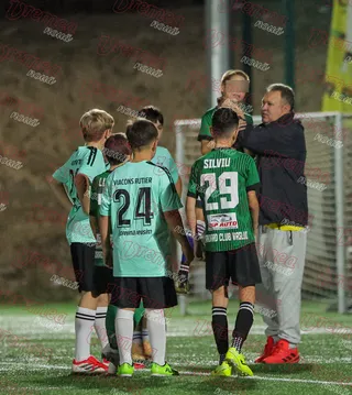 Un grup de copii în echipamente de fotbal verzi și negre stau pe un teren de fotbal lângă un bărbat într-o jachetă închisă la culoare.