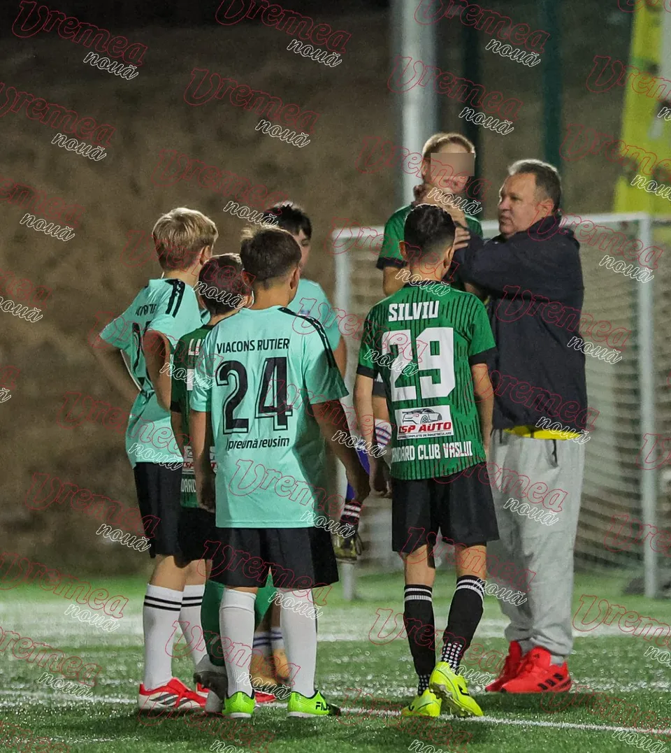 Un grup de copii în echipamente de fotbal verzi și negre stau pe un teren de fotbal lângă un bărbat într-o jachetă închisă la culoare.