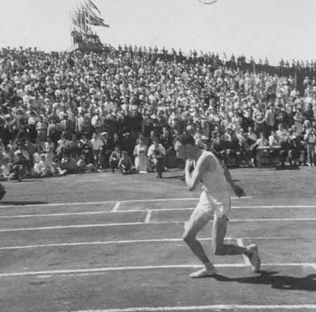 Un bărbat într-un echipament sportiv deschis aleargă pe o pistă de atletism în fața unei tribune pline de oameni.