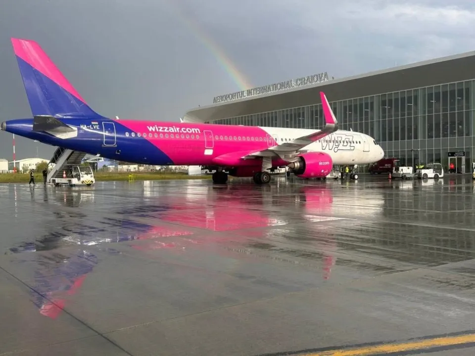 Un avion Airbus de culoare roz și albă stă pe o pistă umedă în fața unui terminal de aeroport sub un arc de curcubeu.