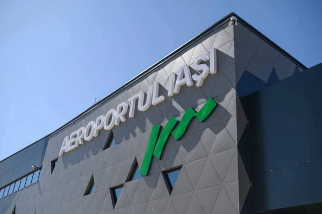 Un semn cu textul AEROPORTUL IAȘI este montat pe fațada unei clădiri gri cu modele geometrice sub un cer senin.