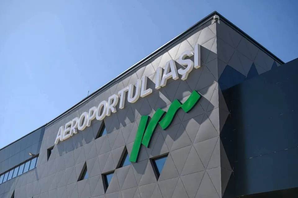 Un semn cu textul AEROPORTUL IAȘI este montat pe fațada unei clădiri gri cu modele geometrice sub un cer senin.