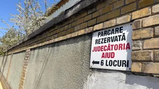 Un semn alb cu text negru și roșu este atașat pe un zid de cărămidă și beton sub un cer senin.