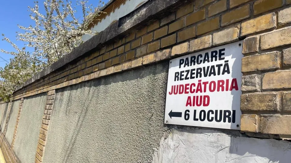 Un semn alb cu text negru și roșu este montat pe un zid de cărămidă și beton sub un cer senin.