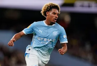 Un bărbat cu părul afro și deschis la culoare poartă un tricou albastru cu logo-uri și aleargă pe un teren de fotbal.