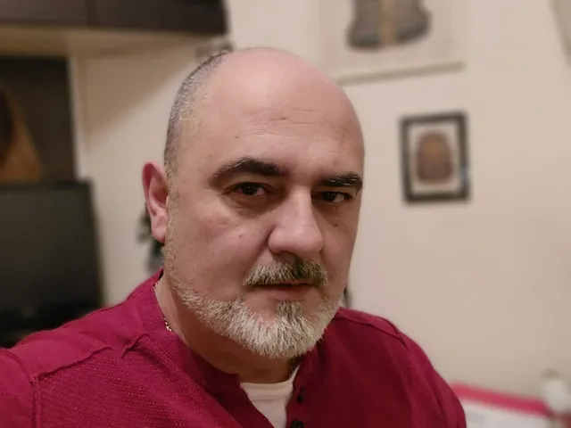 Un bărbat cu barbă și mustață gri, purtând un tricou roșu, privește spre cameră într-un interior.