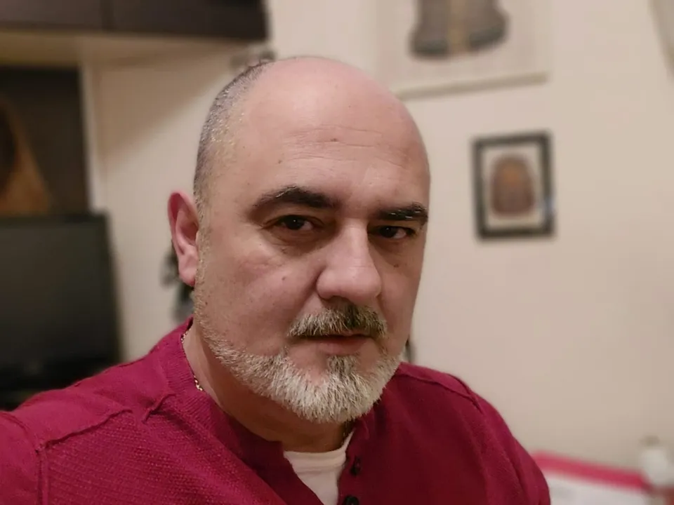 Un bărbat cu barbă și mustață gri, purtând un tricou roșu, privește spre cameră într-un interior.