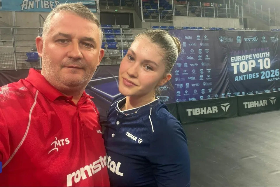 Un bărbat în tricou roșu și o tânără în tricou albastru stau împreună în fața unui panou publicitar de un eveniment sportiv.