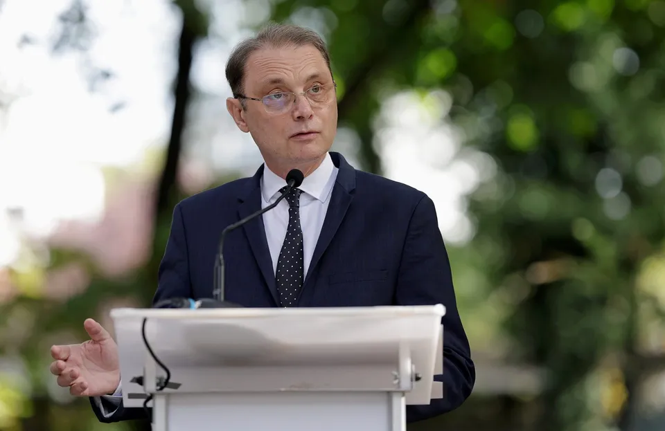 Un bărbat cu ochelari și costum închis stă în spatele unui podium alb sub copaci verzi.