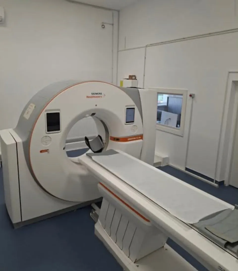 Un aparat medical de tomografie computerizată alb cu detalii portocalii este poziționat într-o încăpere cu pereți albi și podea albastră.