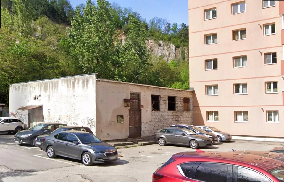 O clădire de un singur etaj cu pereți albi și gri este situată lângă un bloc de apartamente și un teren de parcare cu mașini.