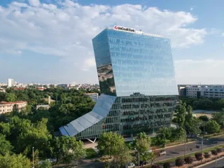 O clădire modernă cu fațadă de sticlă și o structură asimetrică se ridică deasupra unui peisaj urban cu copaci verzi și drumuri.