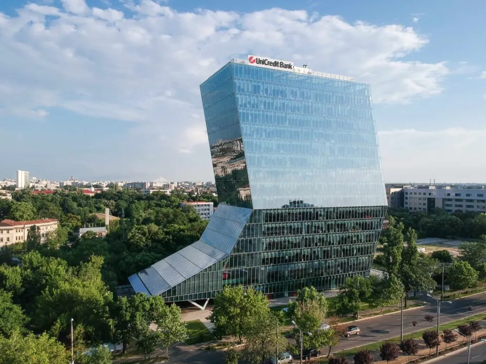 O clădire modernă cu fațadă de sticlă și o structură asimetrică se ridică deasupra unui peisaj urban cu copaci verzi și drumuri.