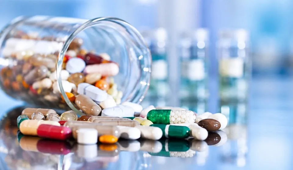 Diverse capsule și tablete colorate sunt împrăștiate pe o suprafață lucioasă lângă un recipient de sticlă răsturnat.