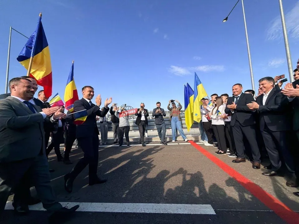 Un grup de bărbați în costume scunde aplaudă pe un drum asfaltat, lângă steaguri și o bandă roșie întinsă pe sol sub un cer senin.