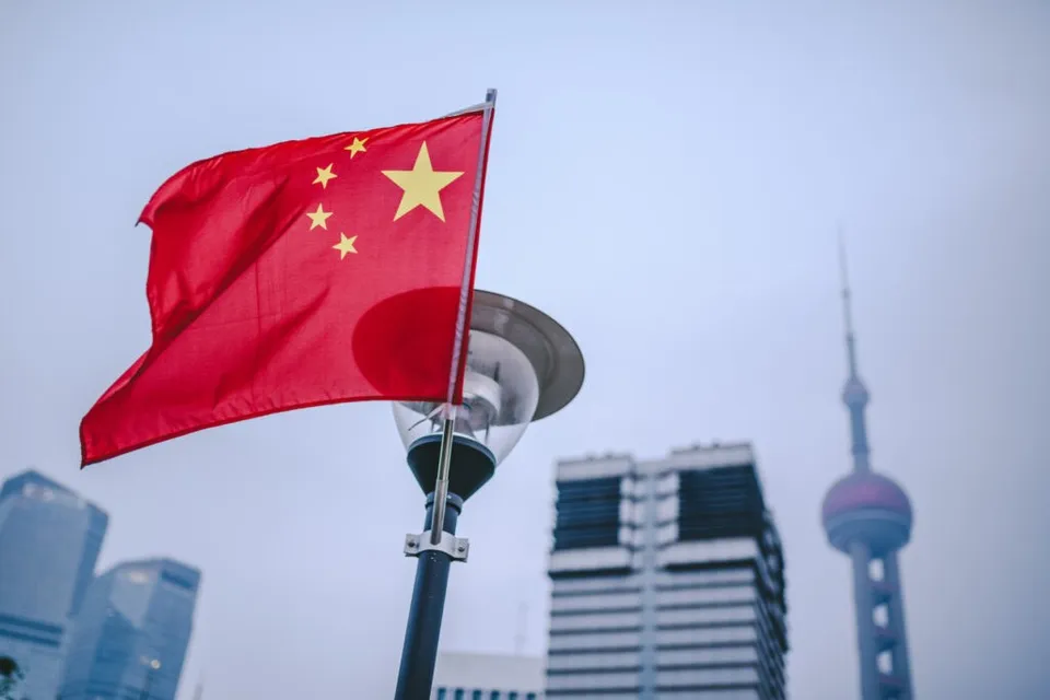 O steag chinezesc roșu cu stele galbene flutură pe un stâlp, având clădiri înalte și un turn în fundal sub un cer închis la culoare.
