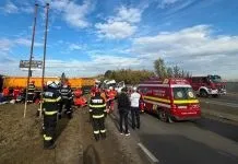 Personal de intervenție în uniforme fluorescente stă lângă un vehicul de urgență roșu și un camion pe o stradă sub un cer înnorat.