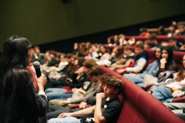 O femeie cu părul lung ține un microfon în mână, privind spre un grup de tineri așezați pe scaune roșii într-o sală de cinema.