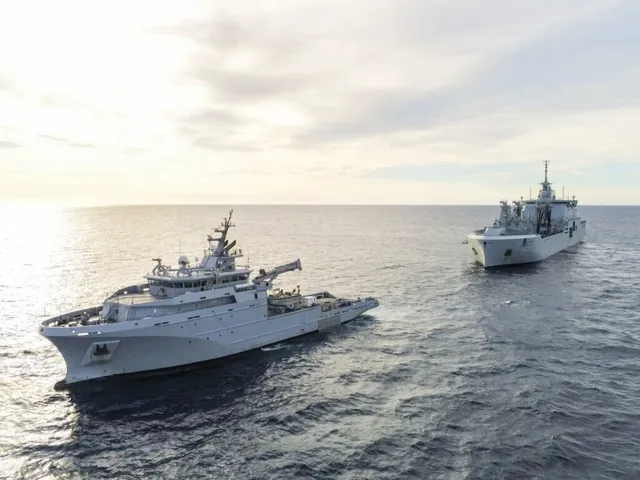 Două nave gri navighează pe o suprafață de apă sub un cer înnorat.