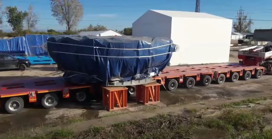 Un utilaj masiv acoperit cu o prelată albastră este transportat pe un trailer lung cu multiple axe pe un teren de pietriș.