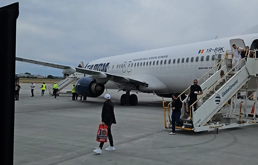 Un avion alb cu inscripții TAROM stă pe o pistă de aeroport, în timp ce pasagerii coboară pe scări mobile lângă fuselaj.