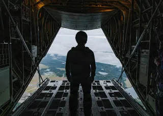 Silueta unei persoane stă în picioare pe rampa deschisă a unui avion, privind spre un peisaj de dealuri și păduri sub un cer înnorat.